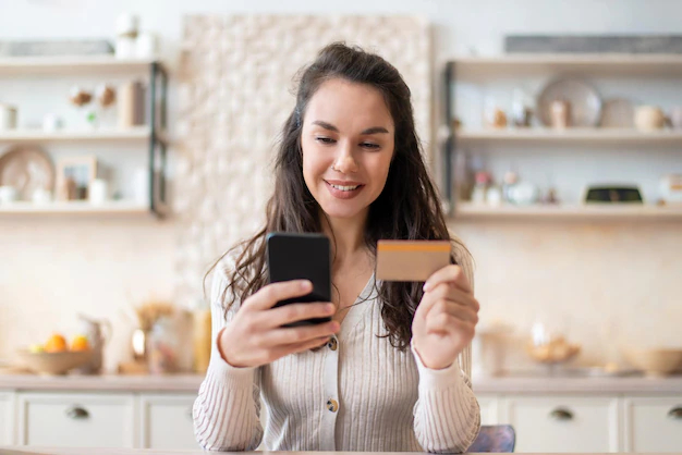 une femme aux expression souriante consultant son smartphone en tenant sa carte de crédit sur fond cuisine flou